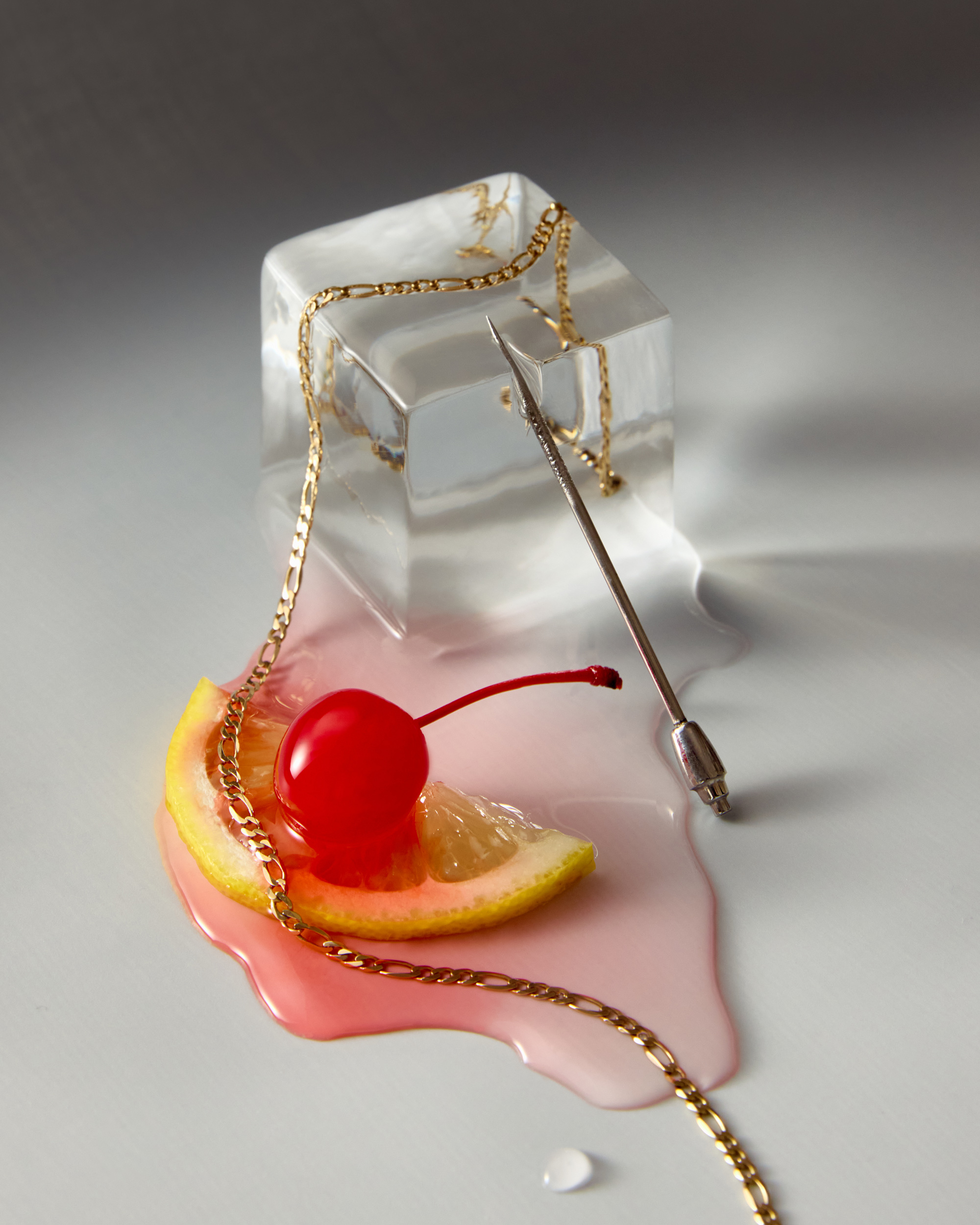 Still life photograph of an ice cube with a gold chain, cocktail spear, lemon slice, cherry, and pink liquid against a gray backdrop.