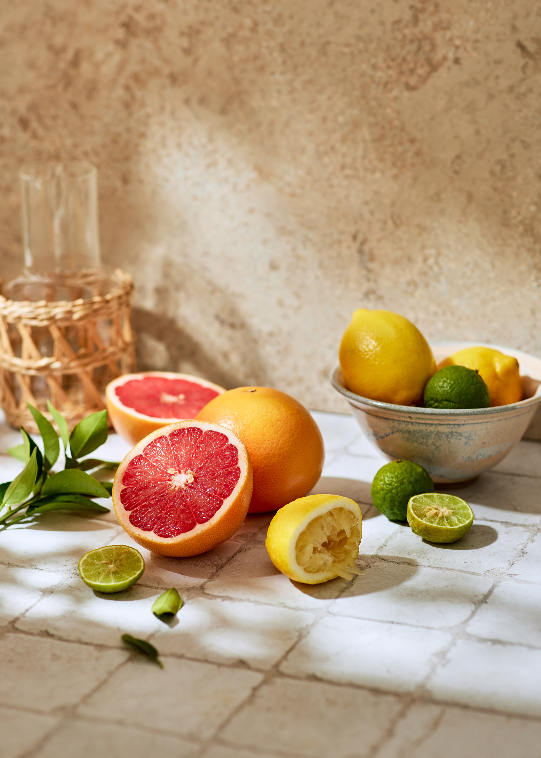 Still life shot of grapefruit, lemons, and limes on a patterned surface, bathed in soft natural light, with a wicker-wrapped vase in the background.