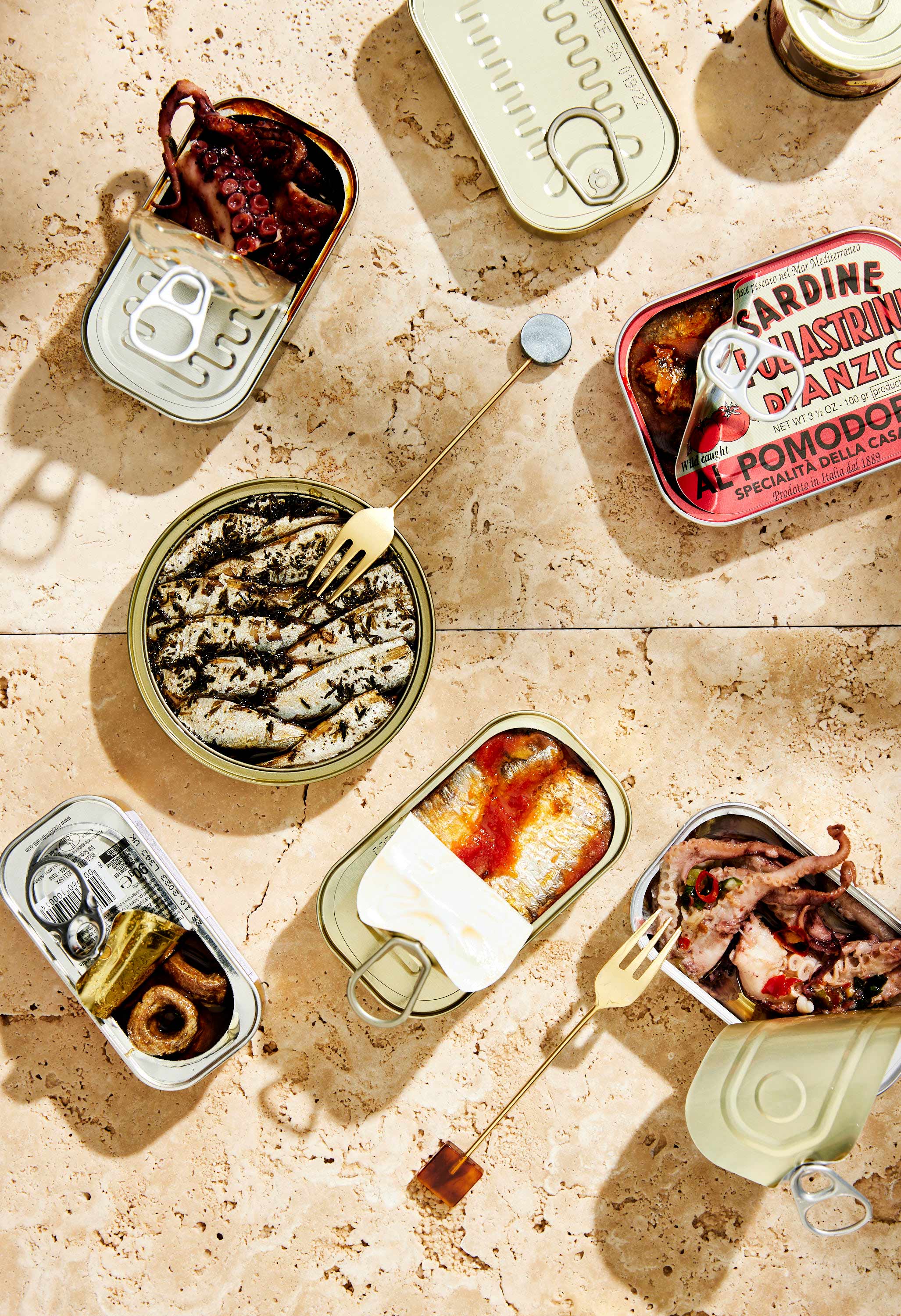 Overhead shot of opened sardine cans on tiled surface, showcasing marinated seafood and condiments in bright, appetizing food photography.