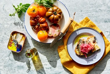 Overhead food photography of heirloom tomatoes, an open sardine tin, and toasted sourdough bread on a grey stone surface