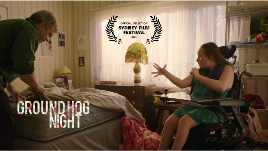 Scene from 'Groundhog Night' film featuring an adult male making a bed and an adult female in a wheelchair in a bedroom.