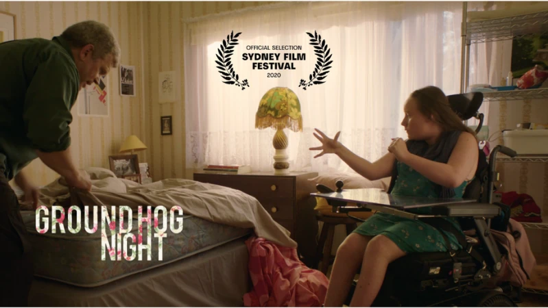 Scene from 'Groundhog Night' film featuring an adult male making a bed and an adult female in a wheelchair in a bedroom.