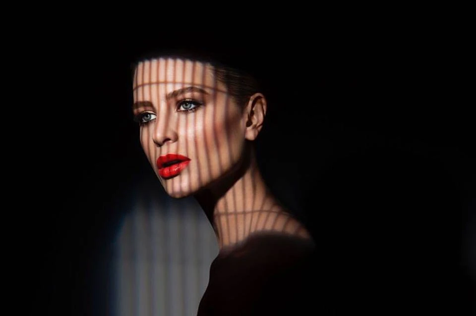Striking beauty portrait of a woman with red lipstick, lit with graphic linear shadows on her face in a dark, artistic studio setting.