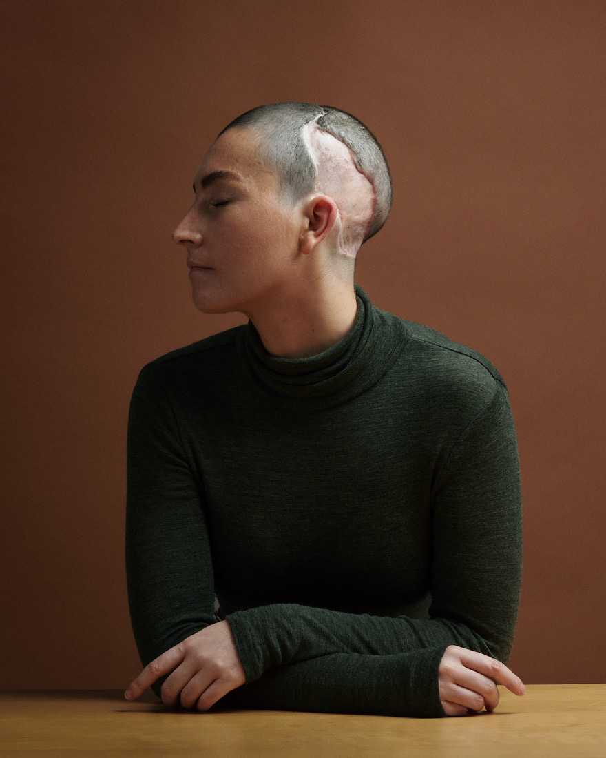 Portrait of an adult female with short hair, eyes closed, and arm resting on a wood table against a brown background.
