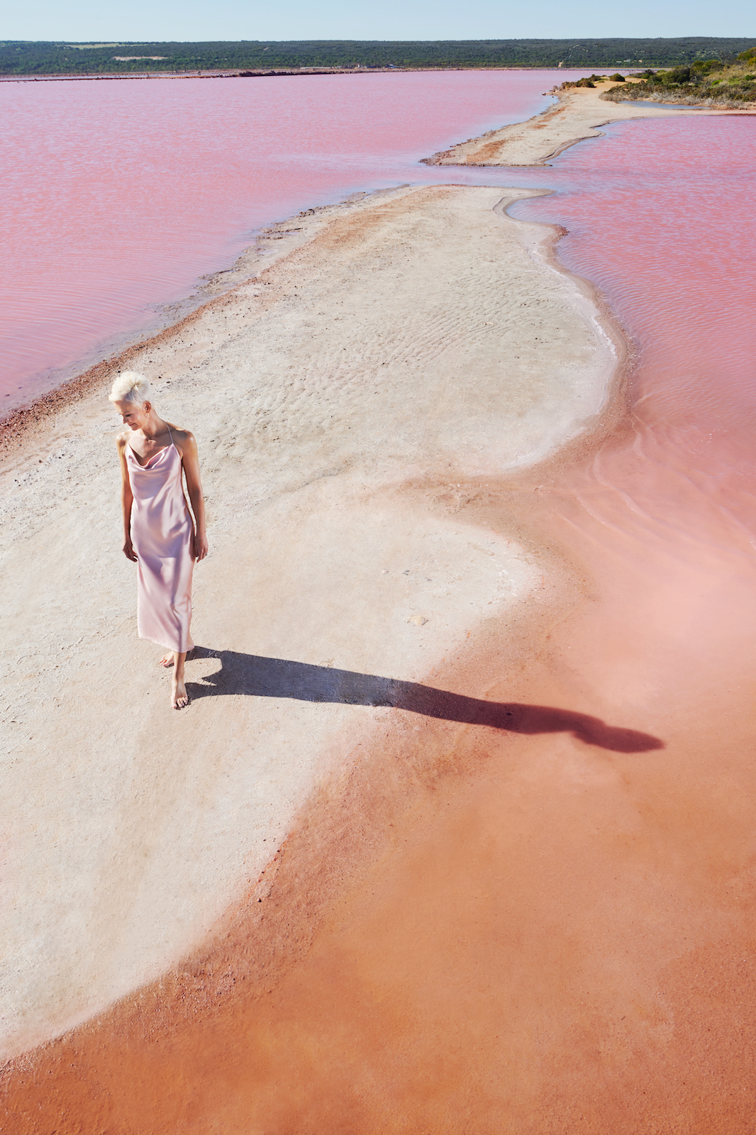 A woman in a light pink dress walks on a white sandbar through a pink lake under a clear blue sky on a bright sunny day.