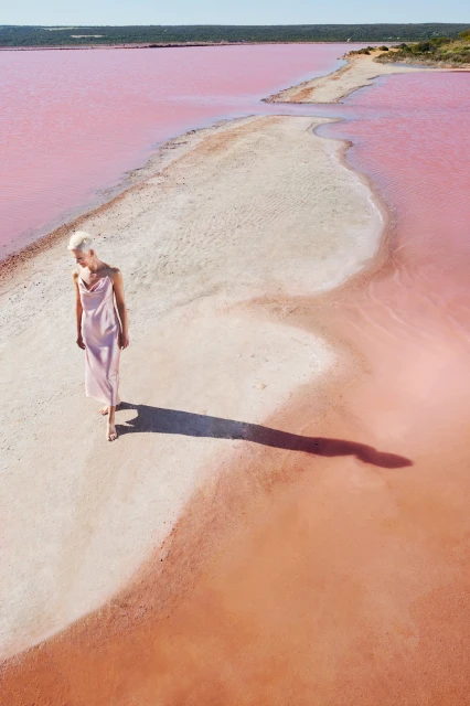 A woman in a light pink dress walks on a white sandbar through a pink lake under a clear blue sky on a bright sunny day.