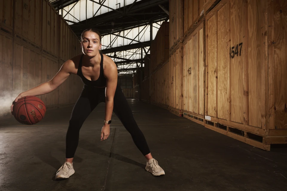 Athletic woman in black sportswear dribbles a basketball in an industrial warehouse-style space with wood paneled walls. 