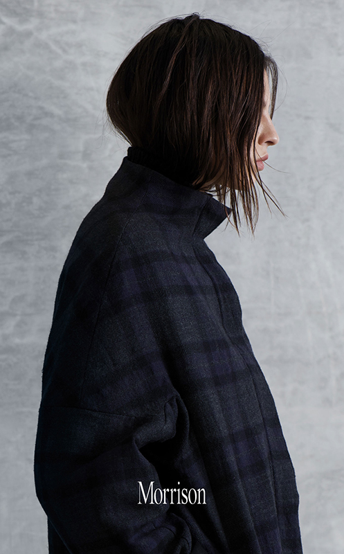 Profile of a woman in a navy plaid jacket against a mottled grey background, showcasing a fashion-forward, sophisticated aesthetic.