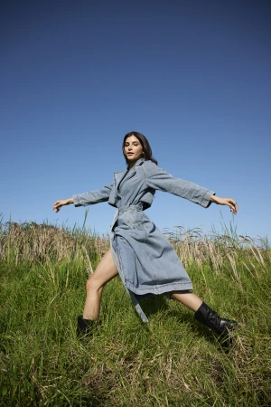 Woman in denim trench coat standing in tall grass under a clear blue sky