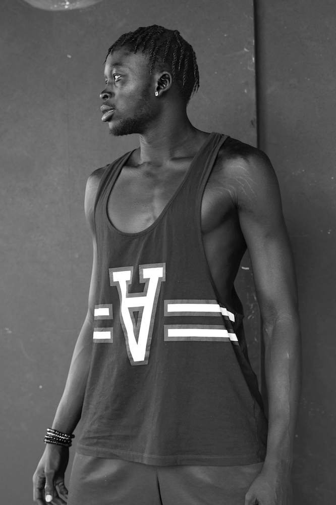 Monochrome portrait of a Black male model in a tank top with the letters 'V' and 'A', posing against a dark grey wall with a serious expression.