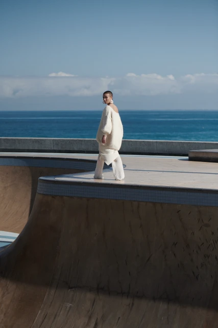 A slender woman in a cream-colored sweater and white pants walks on a skatepark ramp under a bright blue sky with the ocean visible.