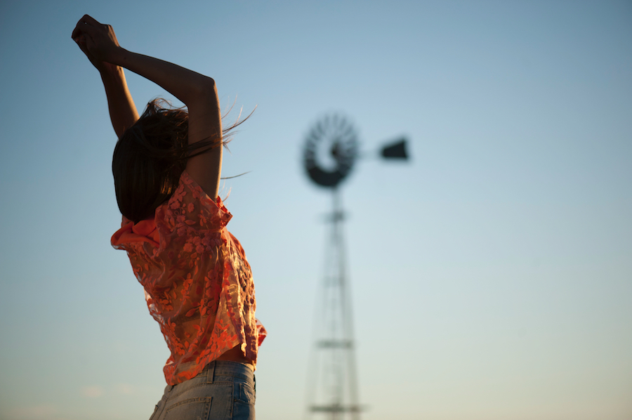 A woman stretches arms against a blue sky, with a blurred windmill in the background, creating a serene and expansive outdoor scene.