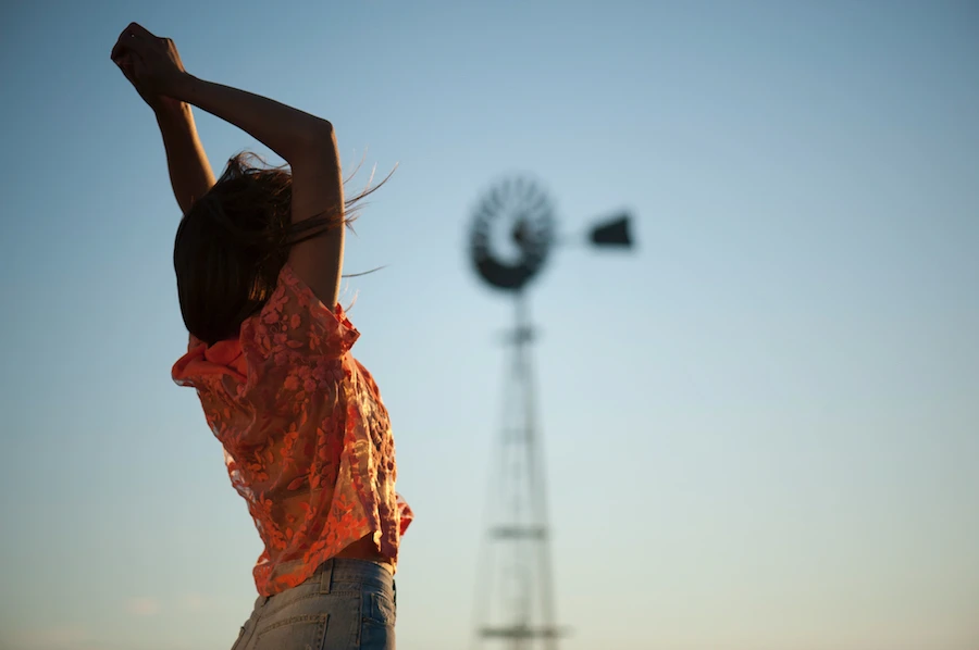 A woman stretches arms against a blue sky, with a blurred windmill in the background, creating a serene and expansive outdoor scene.