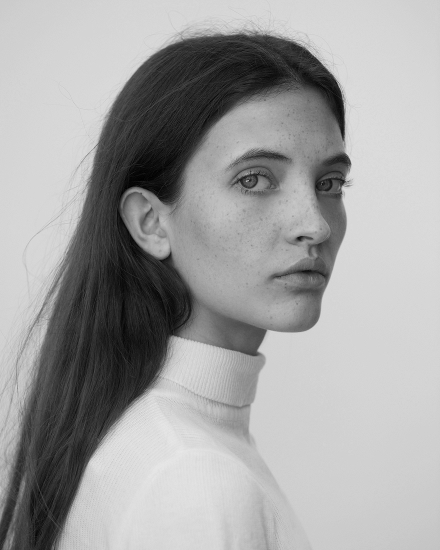 Portrait of a young adult woman with long dark hair looking over her shoulder wearing a white turtleneck against a plain white background.