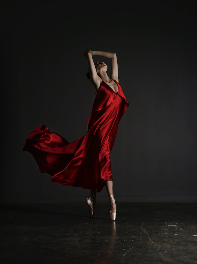 Ballet dancer poses in a red dress, en pointe, with arms raised overhead in a dark photo studio showcasing elegance and motion.