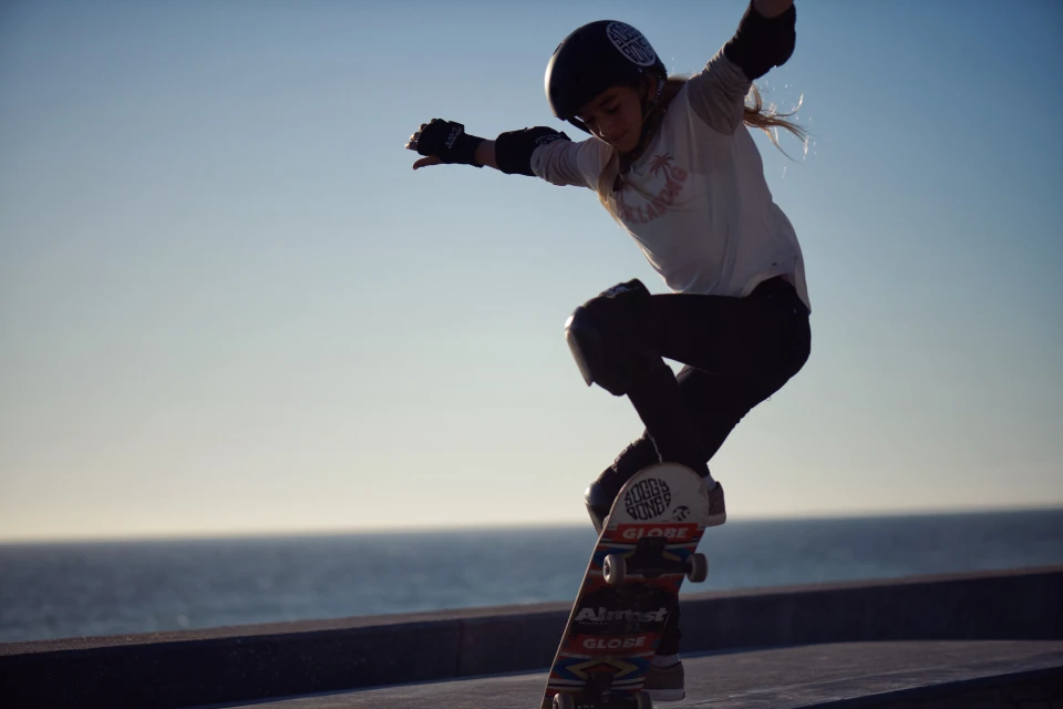 A young female skateboarder in mid-air wearing safety gear against a blue sky and ocean backdrop performs a stunt with her skateboard.