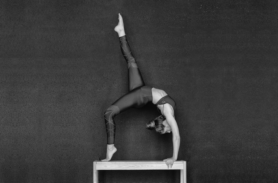 A woman in athletic wear is doing a backbend and lifting one leg up in the air while balancing on a wooden structure in black and white.