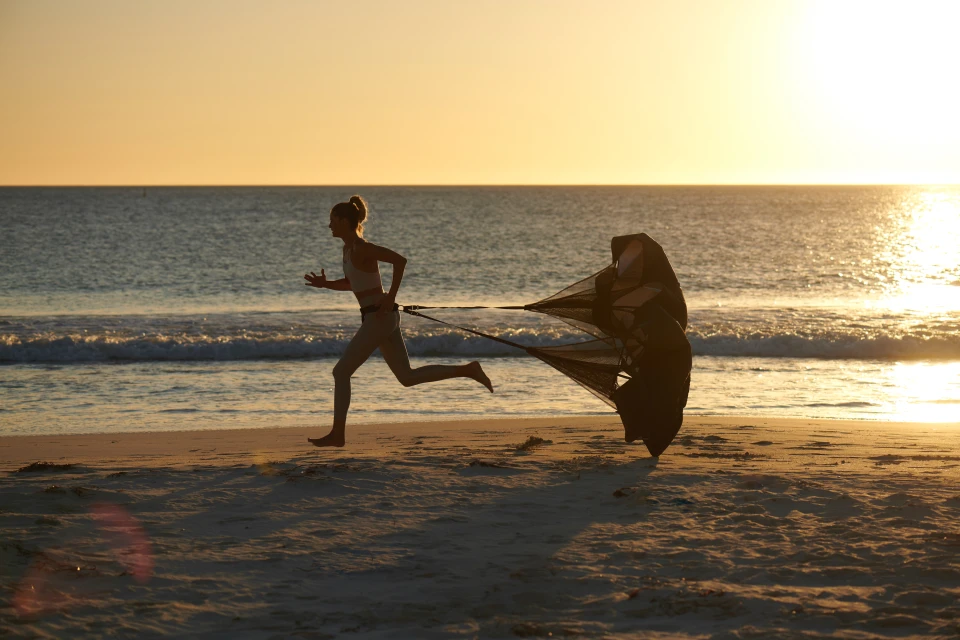 A woman exercises on a sandy beach at sunset, running and pulling a resistance parachute, with ocean waves and a bright sun.