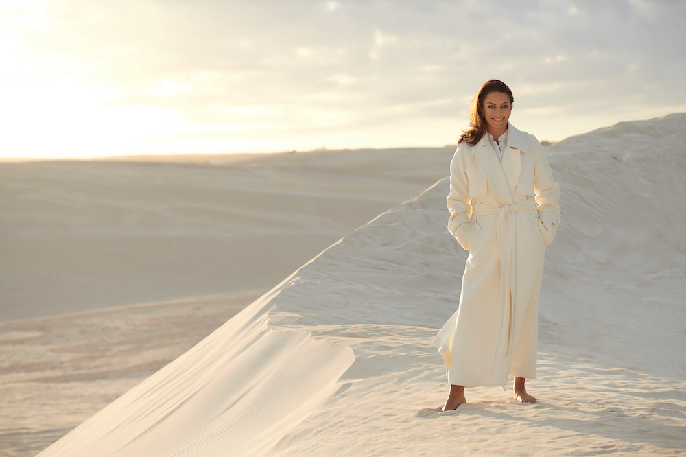 Adult woman in a white trench coat standing on sand dunes in desert landscape with a sunny sky, fashion shot, beauty shot, travel
