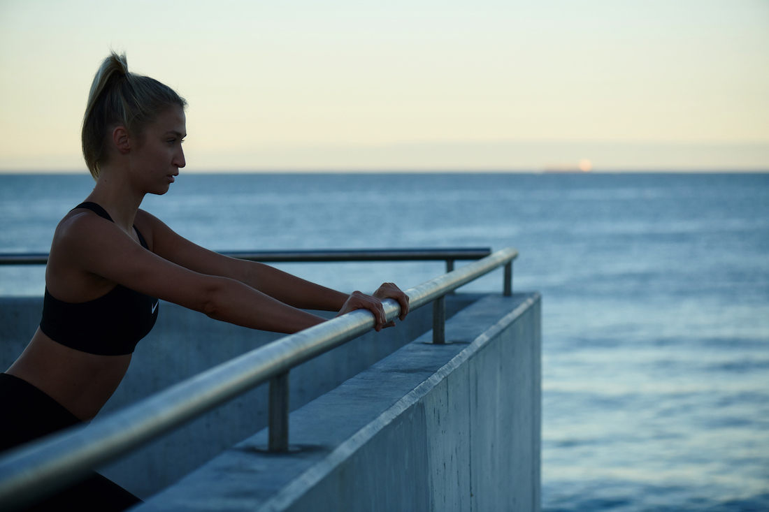 Athletic blonde woman stretching arms on handrail, dressed in activewear, with ocean and sky in the background, at dusk or dawn.