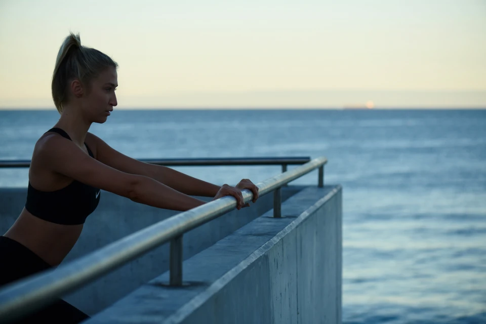Athletic blonde woman stretching arms on handrail, dressed in activewear, with ocean and sky in the background, at dusk or dawn.