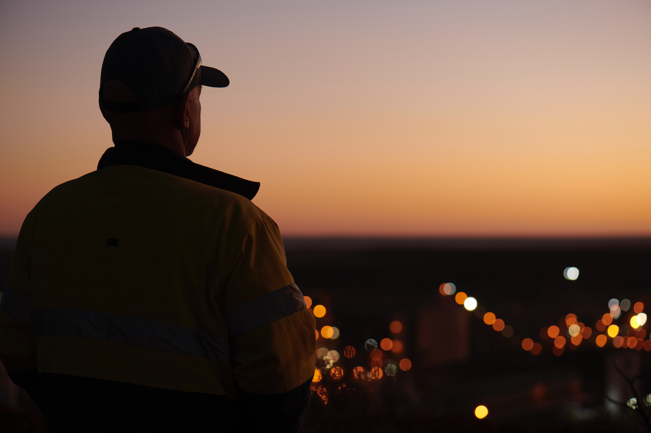 Person in workwear gazes at cityscape at sunset, back view. Protective outerwear, cap, dramatic gradient sky, and bokeh lights.