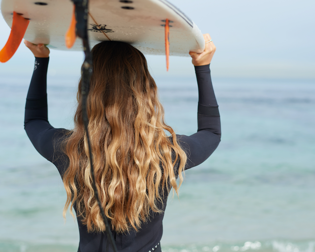Back view of a woman carrying a surfboard on her head at the beach with a view of the ocean. She has long wavy blonde hair.