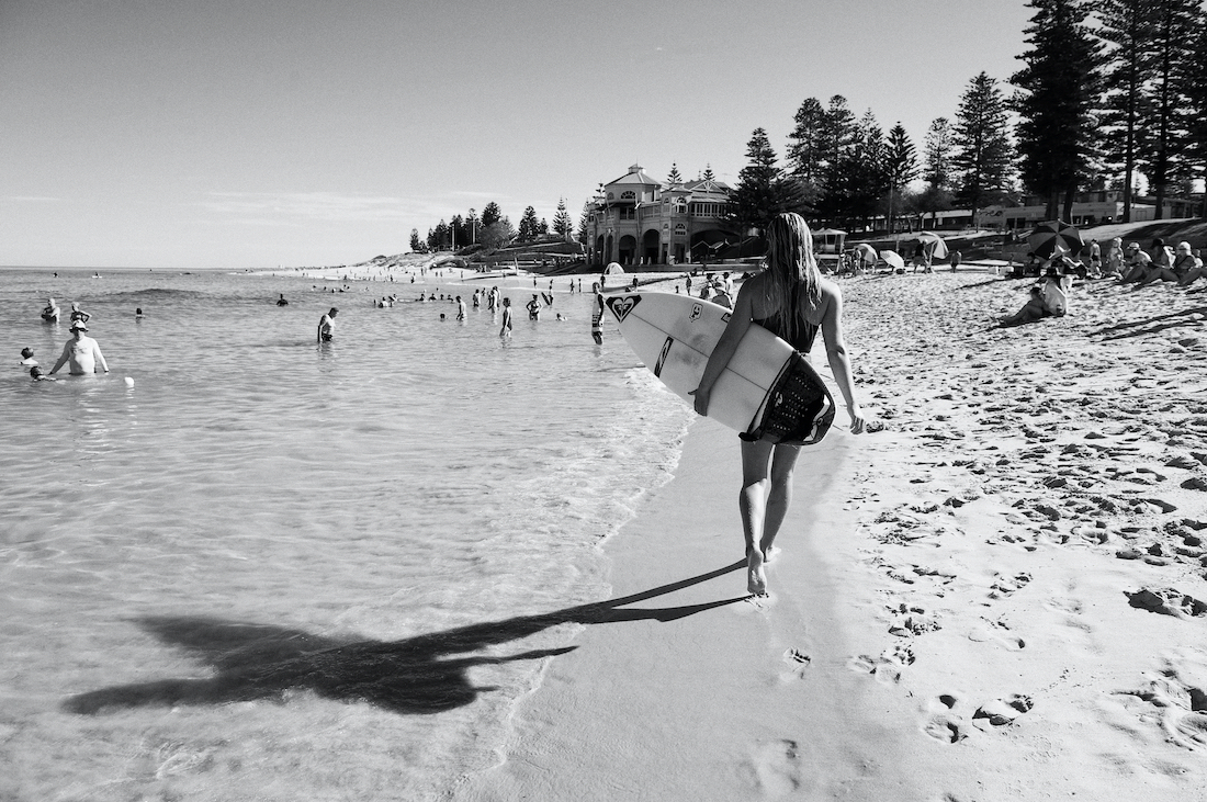 Black and white shot of a woman walking on the beach with a surfboard, ocean and many people in the background on a sunny day.