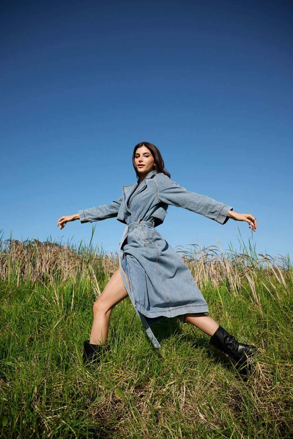 Woman in denim trench coat standing in tall grass under a clear blue sky