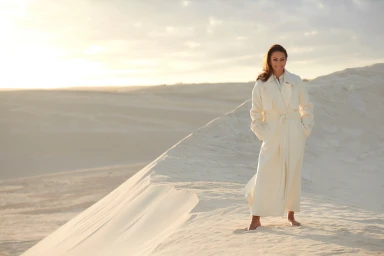 Woman in a long white coat standing barefoot on a sandy dune at sunset by Stef King