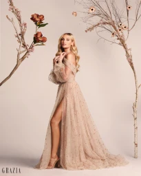 Blonde woman in an off-the-shoulder sheer embroidered gown with a high slit standing against a seamless background with decorative branches.