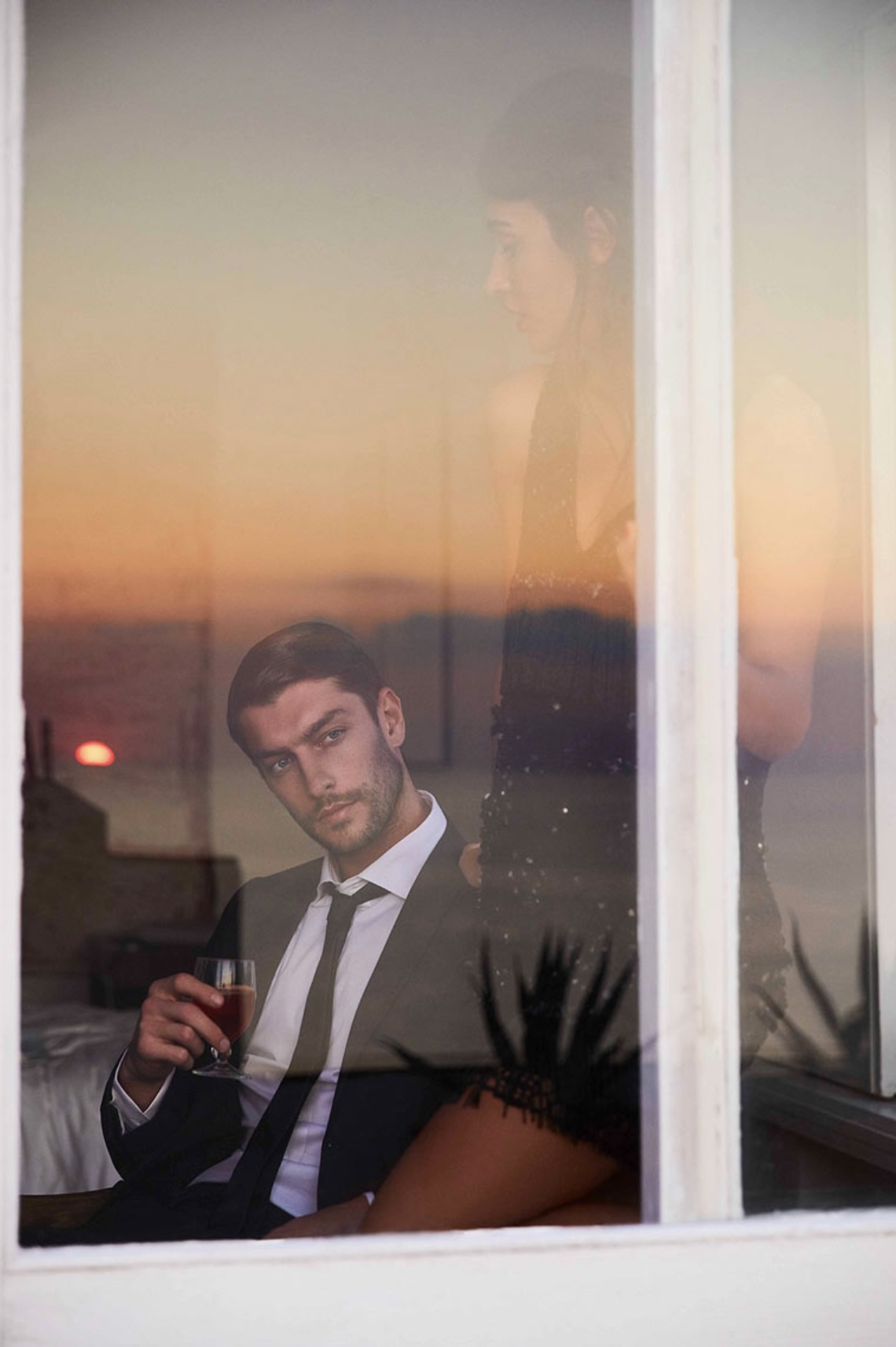Man in suit holds drink by window, woman in background, sunset view. Evening fashion photography. Indoor hotel setting with moody lighting.