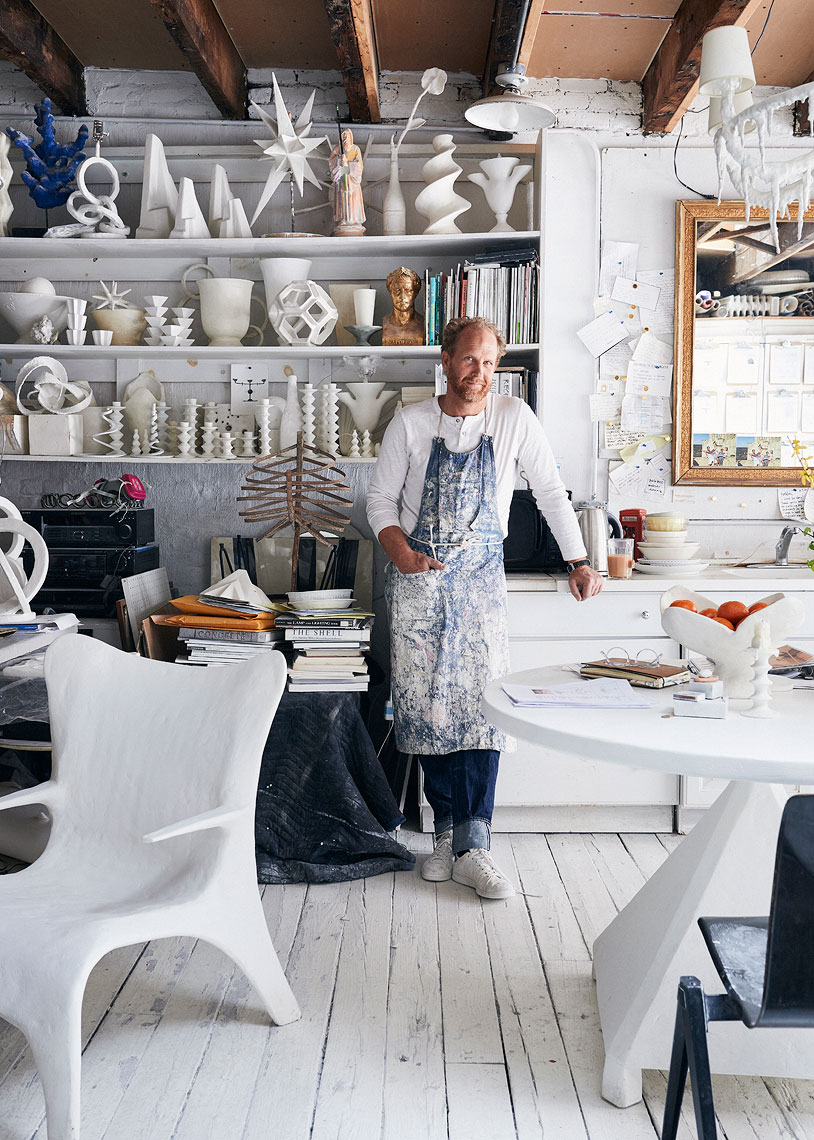 Man with fair skin standing inside a bright art studio space, white sculptures on shelving, white walls, and a white painted floor.