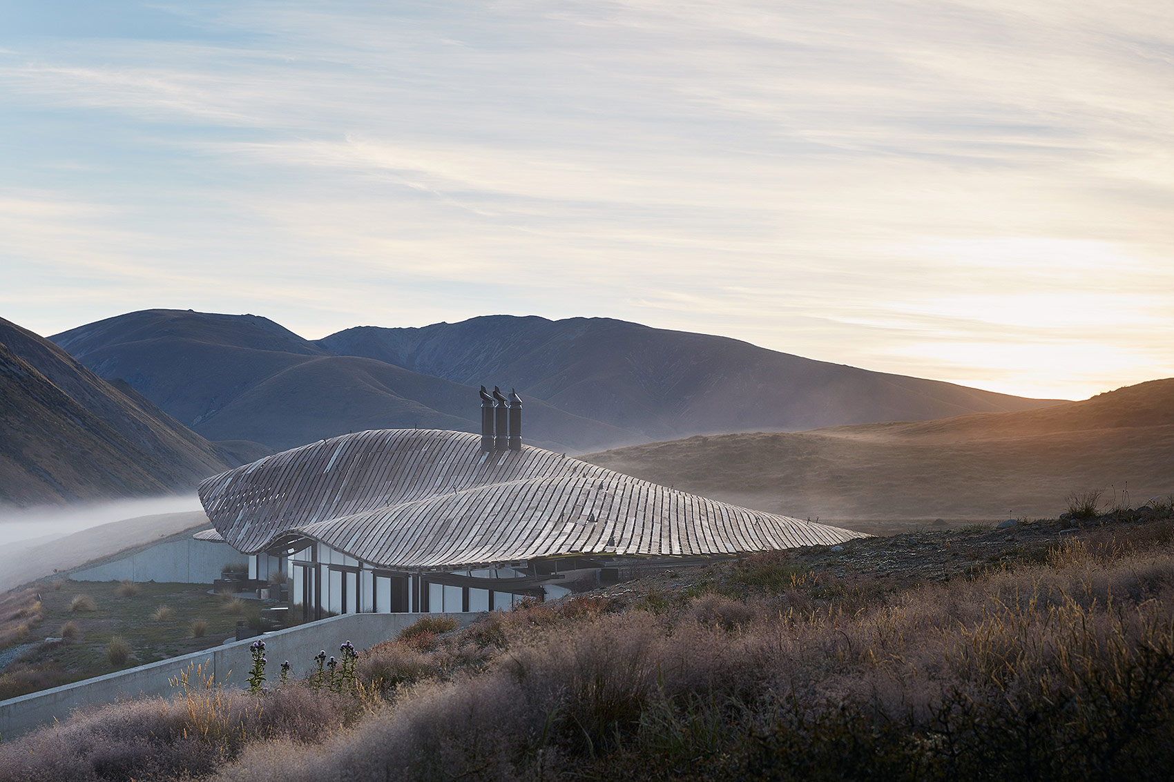 Architecture of a modern house with a curved roof nestled in a valley with mountains, illuminated by the soft glow of sunrise.