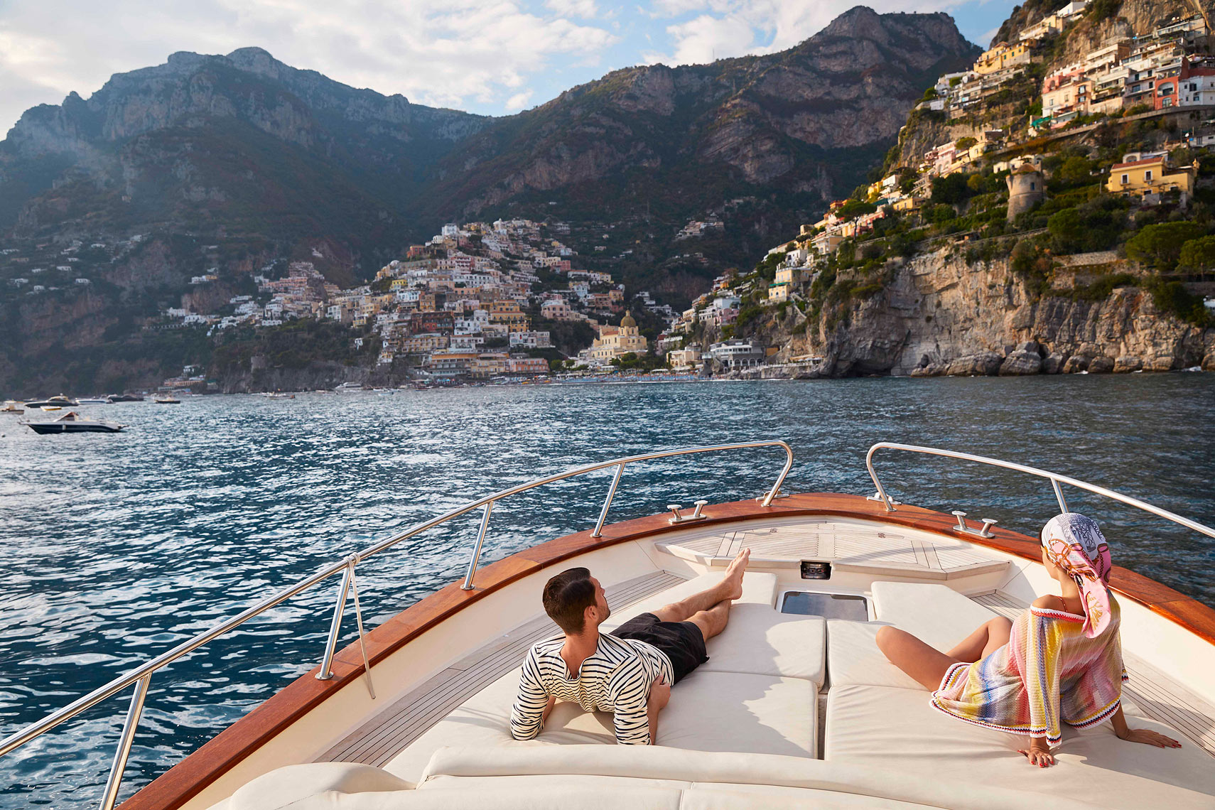A couple relaxes on a luxury motor yacht with a coastal Italian town backdrop, featuring blue water and clear skies on a sunny day.