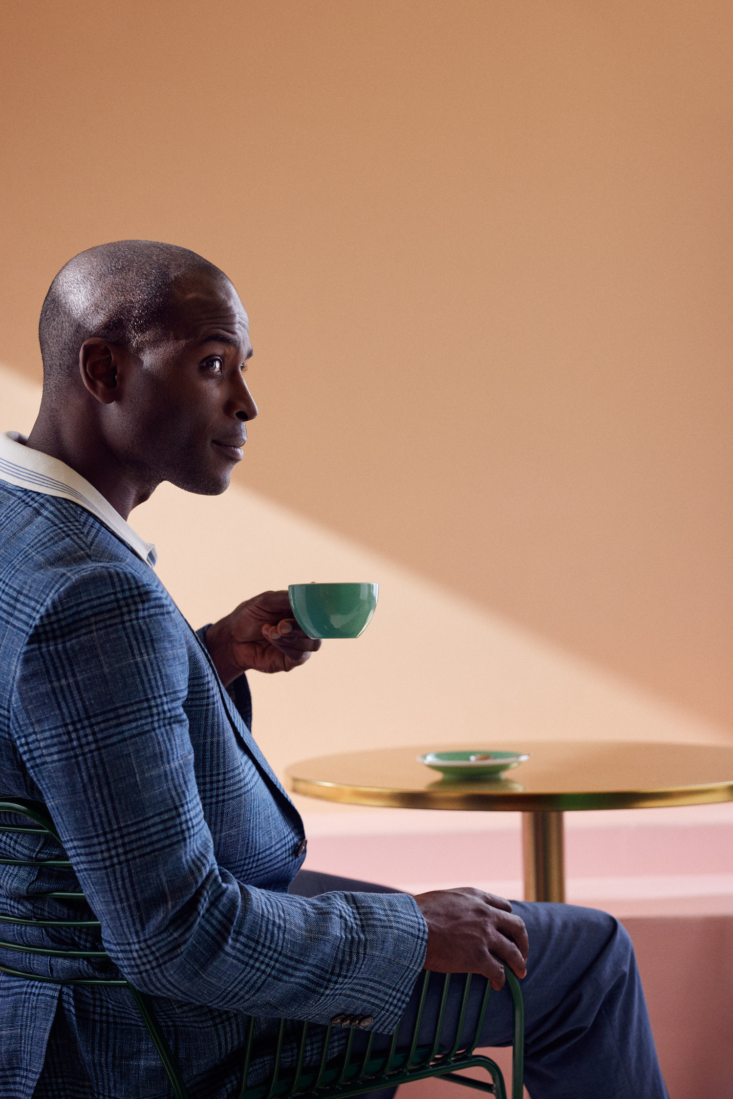 Dark-skinned adult male seated, wearing a plaid jacket, holding a green teacup at a gold table in a neutral-toned room. 