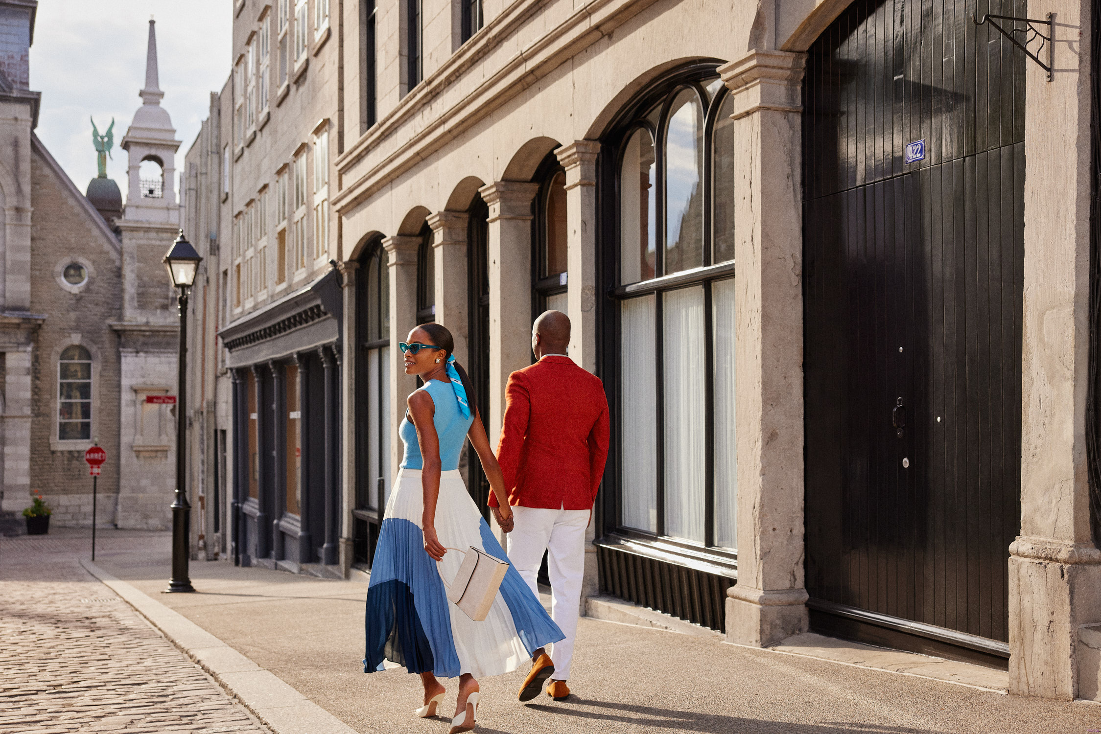 Couple walking down a cobblestone street in an urban setting; the woman wears a blue top and pleated skirt, and the man a red jacket.