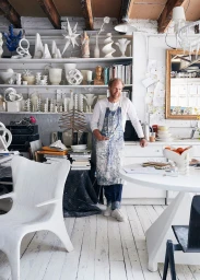 Man wearing a paint-splattered apron standing in a white artist studio filled with sculptures and shelves