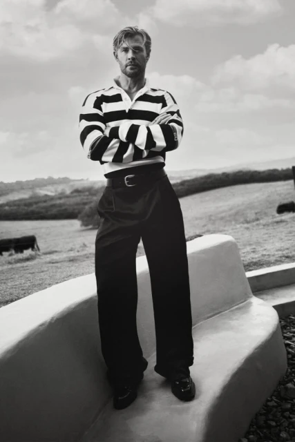 Black and white portrait of an adult male in a striped shirt and wide-leg pants posing on a concrete structure outdoors in a rural landscape.