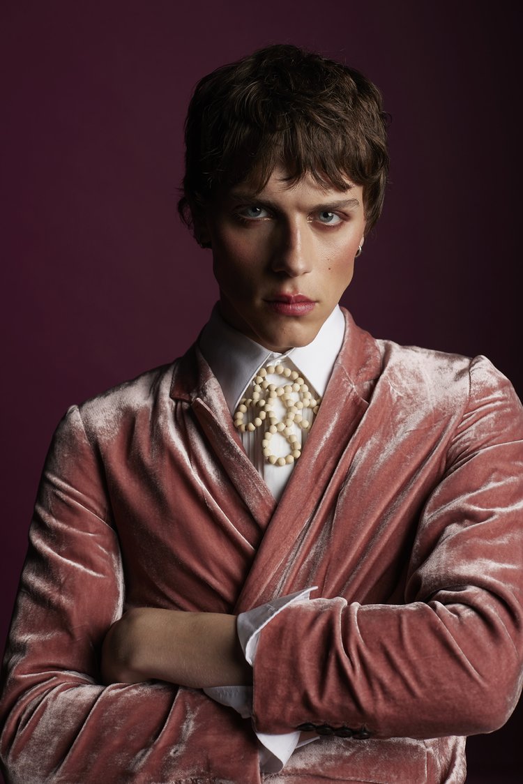 Headshot of a male fashion model in a pink velvet blazer and white shirt with crossed arms against a dark purple studio backdrop.