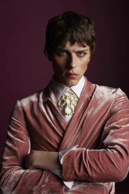 Headshot of a male fashion model in a pink velvet blazer and white shirt with crossed arms against a dark purple studio backdrop.