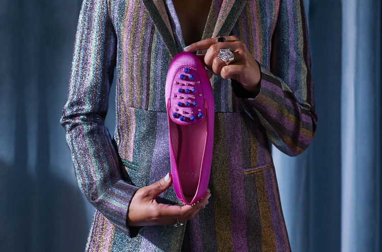 Person holding a pink satin ballet flat with blue embellishments, wearing a sparkling striped blazer and a statement ring against a blue backdrop.