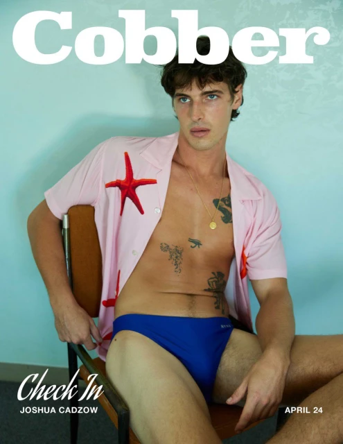 Male model sits in chair, wearing blue swim brief and open pink shirt decorated with red starfish in a studio environment.