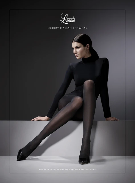 Elegant woman in black tights poses gracefully against a dark background in a fashion advertisement, showcasing luxury Italian legwear.