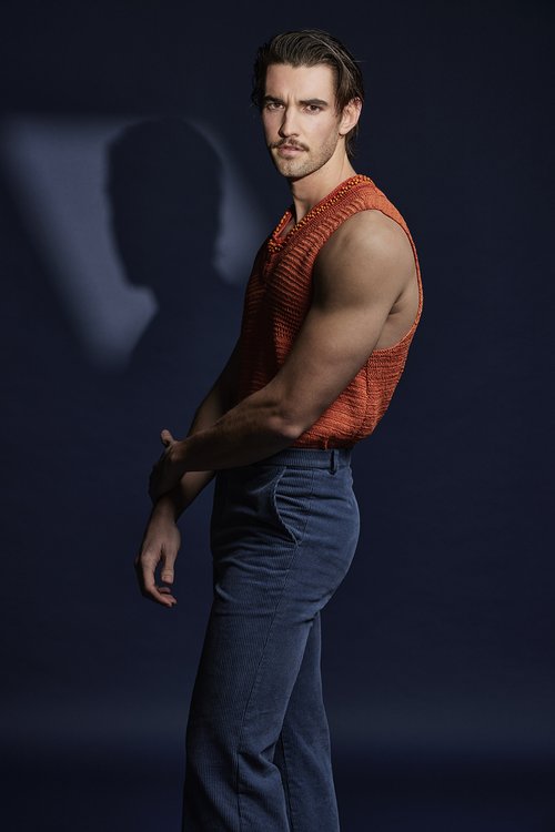 Studio portrait of a young adult man in an orange knit vest and blue jeans posing against a dark blue background with soft studio lighting.