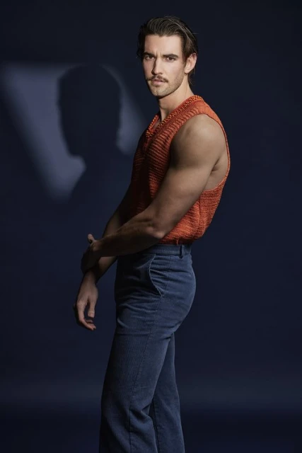 Studio portrait of a young adult man in an orange knit vest and blue jeans posing against a dark blue background with soft studio lighting.