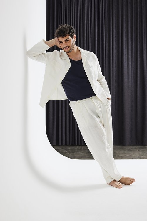 Man in white linen suit leans against a white architectural wall, posing in front of a dark curtain backdrop with a confident expression.