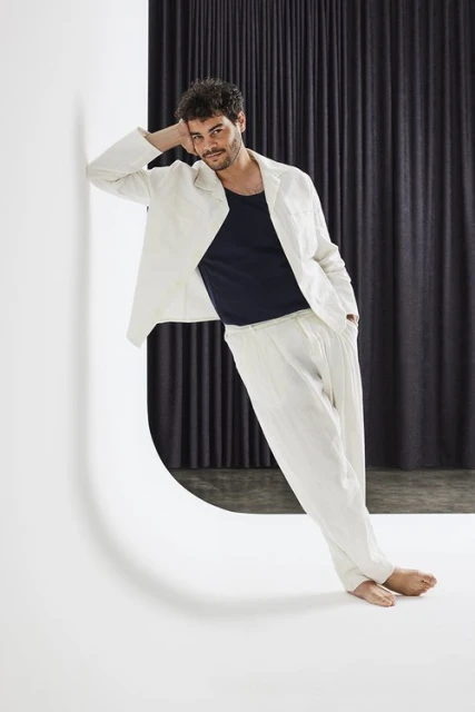 Man in white linen suit leans against a white architectural wall, posing in front of a dark curtain backdrop with a confident expression.