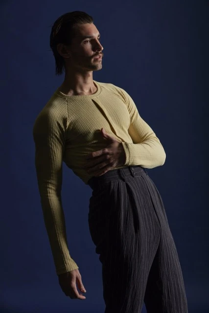 Portrait of a male fashion model with dark hair and a mustache in a yellow long-sleeve shirt and pinstripe pants against a blue backdrop.