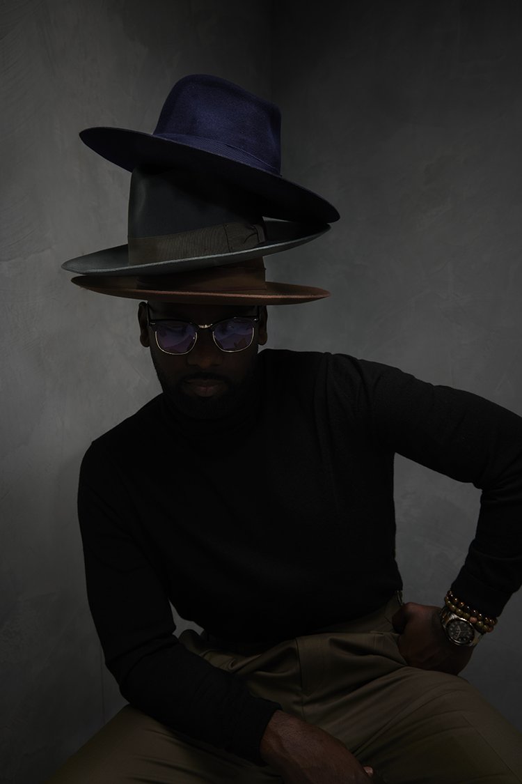 Dark-skinned man poses in long-sleeve shirt and slacks with sunglasses and three hats stacked on his head against a gray backdrop.